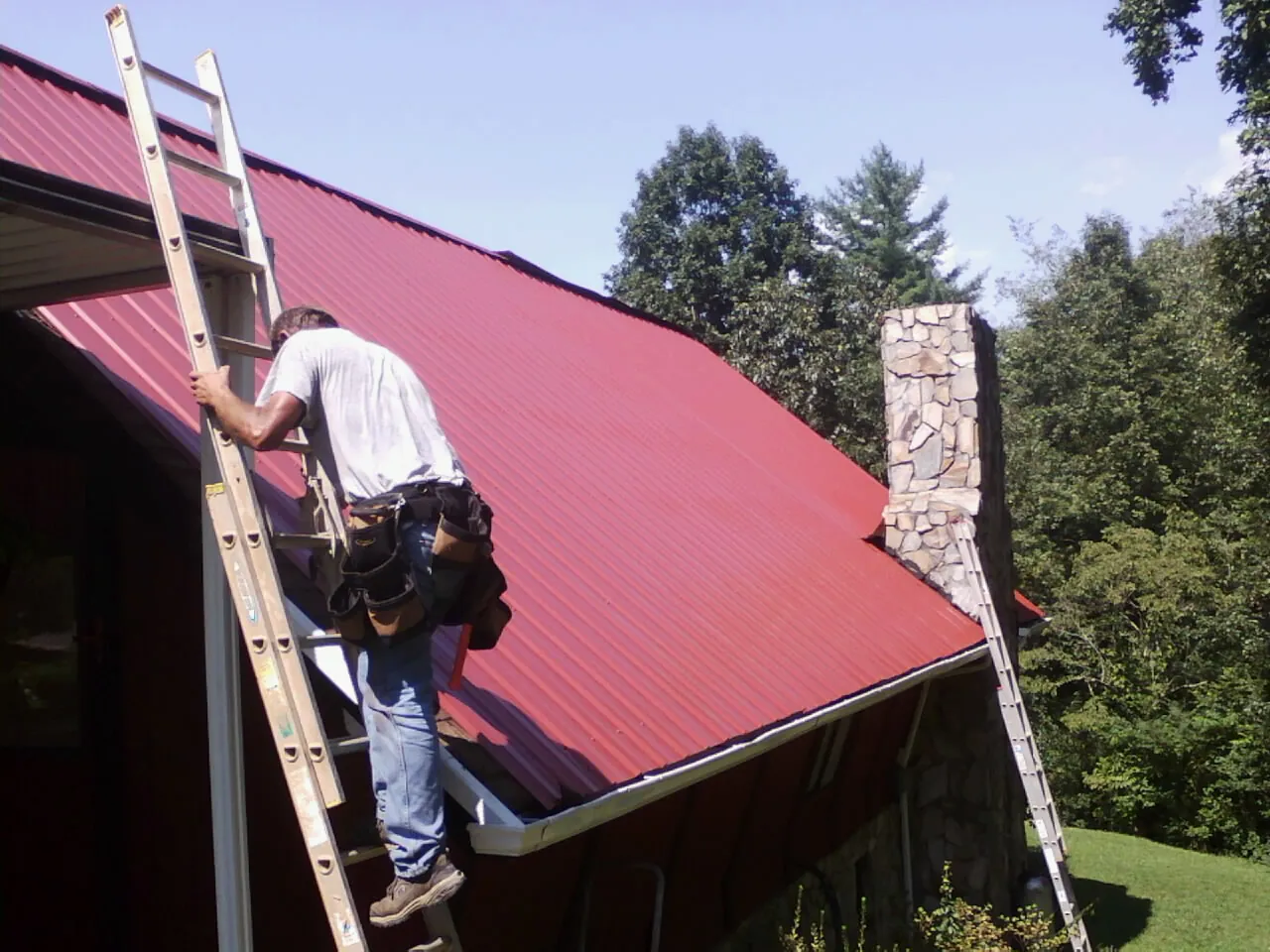 Roofer on a ladder beside a red metal roof and stone chimney — Soffit & Fascia Repair by Roofing Brothers in Missouri City